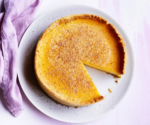 Plate of egg custard tart with a slice missing, topped with ground nutmeg, on a light surface near a purple cloth.