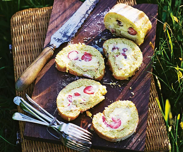 "Strawberry and cream Swiss roll slices on a wooden board with a knife and forks on the side."