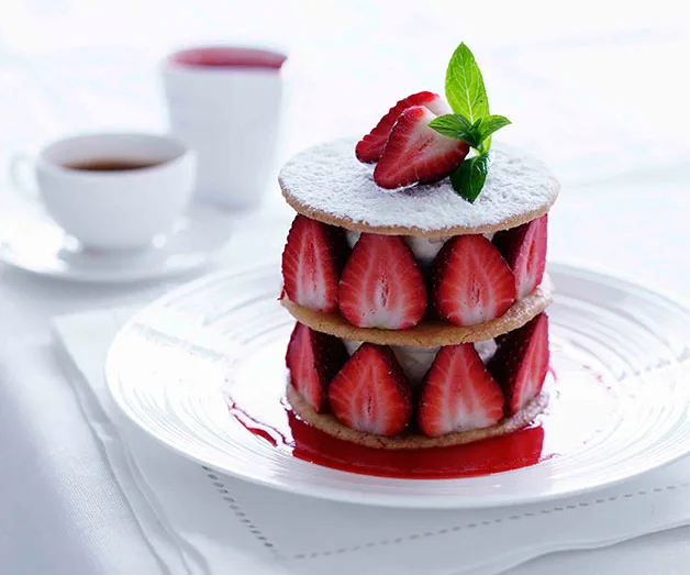 Layered dessert with strawberries, cream, and biscuits on a white plate, topped with mint and powdered sugar.