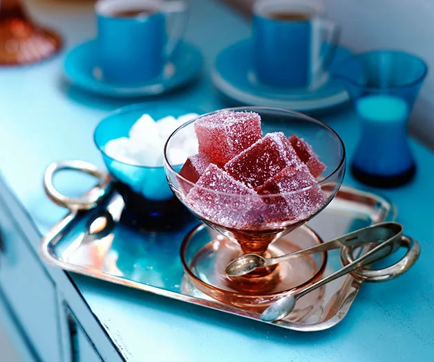 Glass of red sugared jellies on tray with two spoons; blue cups and sugar cubes in background.