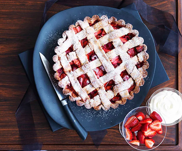 Strawberry pie with lattice crust, dusted with sugar, on black plate with a knife, bowl of cream, and sliced strawberries.
