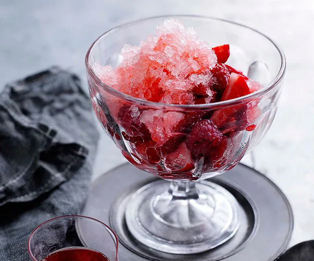 Glass bowl with red fruit salad topped with pink crushed ice on a silver plate.