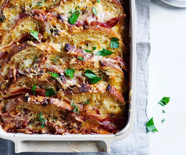 Savory bread and butter pudding with bacon, cheese, herbs in a baking dish on a light tablecloth.
