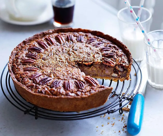 Pecan pie on cooling rack with a slice removed, next to glass of milk with straws and a drink in the background.