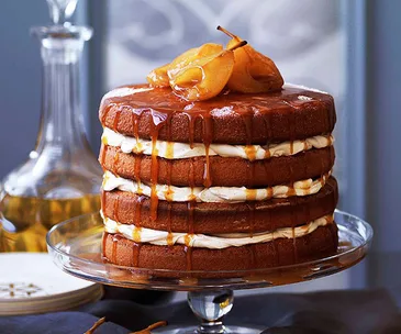 Photo of glass cake stand with layered brown sponge cake with white cream filling and drippiong caramel, topped with poached pears