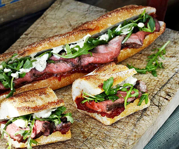 A roast beef sandwich with arugula and sauce on a wooden board, presented in a baguette and cut into slices.