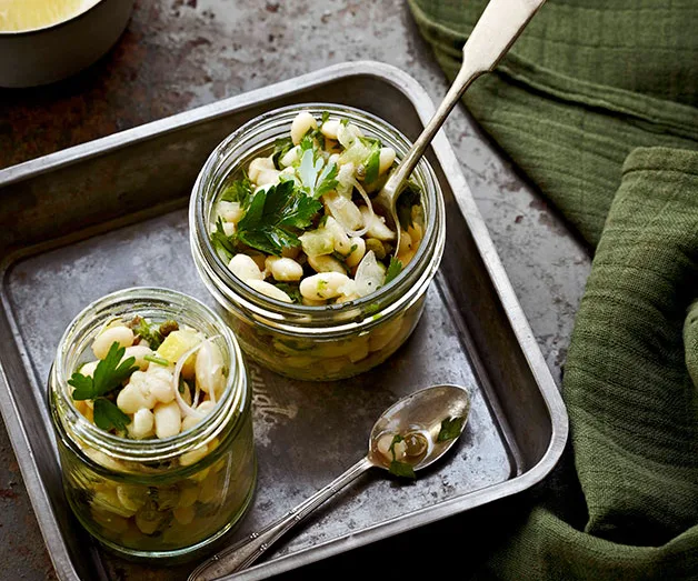 Two jars of marinated white beans with herbs and onions on a metal tray, with a spoon beside.