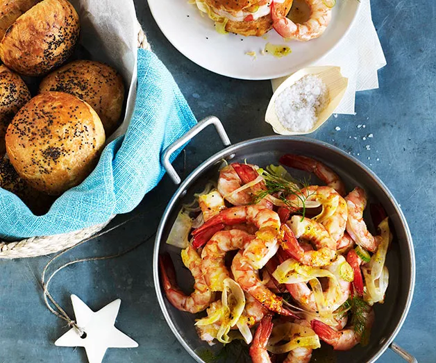 A bowl of cooked prawns with fennel and herbs, next to a basket of poppy seed rolls on a blue table.