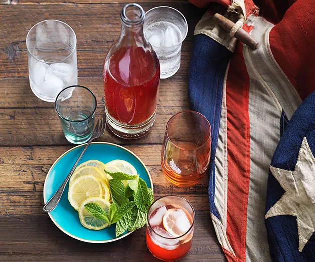 A bottle and glasses with red drink, ice, and lemonade on a wooden table with mint and a vintage American flag.