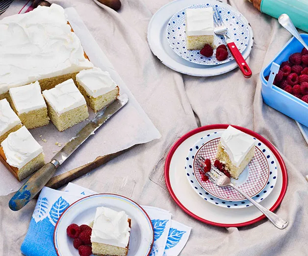 Squares of frosted cake on plates with raspberries and forks on a cloth.