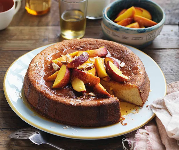 A moist almond cake topped with sliced peaches and syrup is served on a plate with a slice missing, surrounded by drinks.