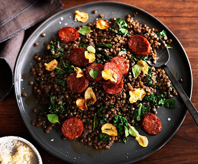 Plate of lentils with chorizo, kale, and garlic chips on a dark wooden table, garnished with herbs.