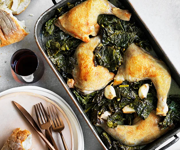 Roasted chicken legs with garlic and leafy greens in a baking dish; bread and wine on the side.