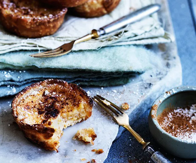 A close-up of a partly eaten pastel de nata on a marble surface with a small fork and sprinkled cinnamon nearby.