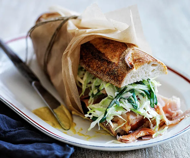 Sandwich wrapped in paper with ham and slaw on a plate, beside a knife with mustard.