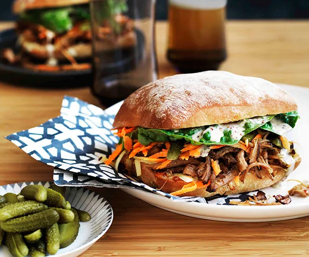 Pulled pork sandwich with lettuce and carrots on a ciabatta roll, served with pickles, glass, and beverage in background.
