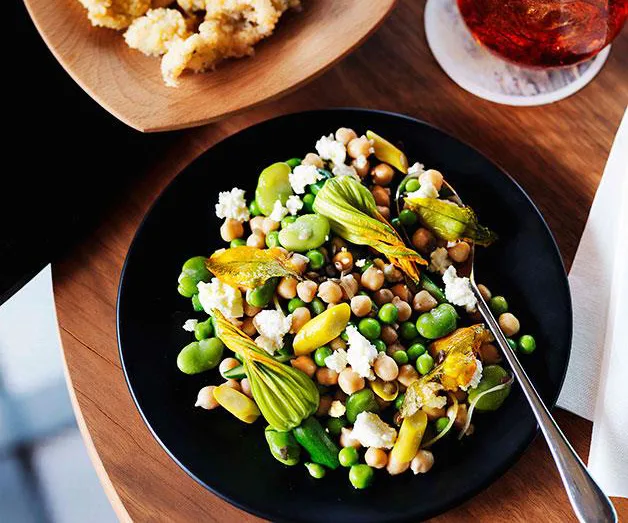 Chickpea, broad bean, and zucchini blossom salad with ricotta on a black plate on a wooden table.