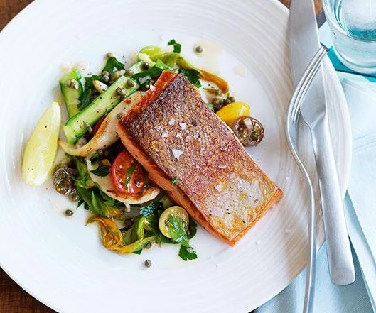 Grilled salmon fillet on a white plate with colorful vegetables and greens, garnished with capers next to cutlery.