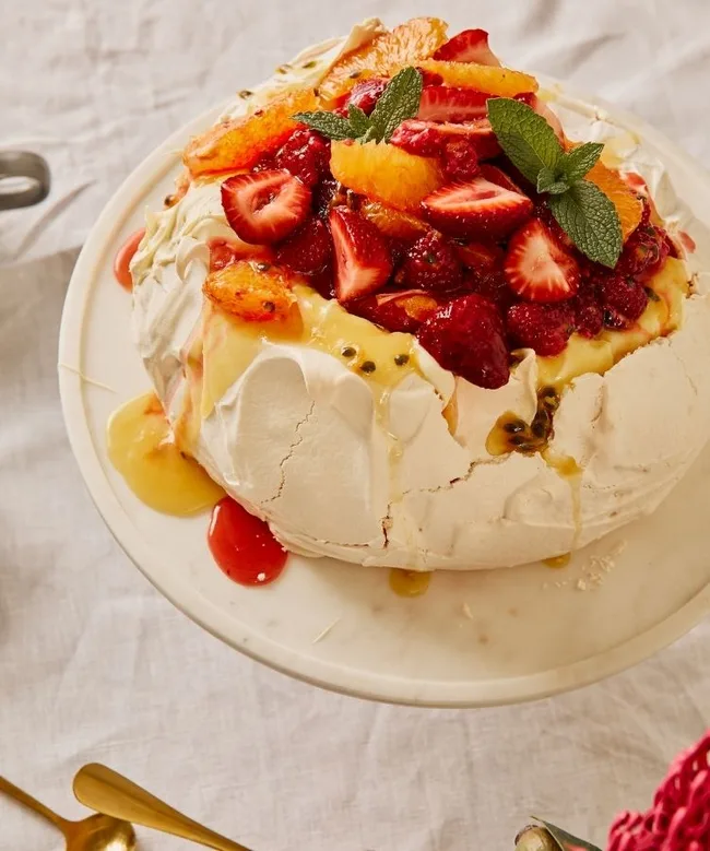 A pavlova topped with strawberries, orange slices, and mint on a white plate.