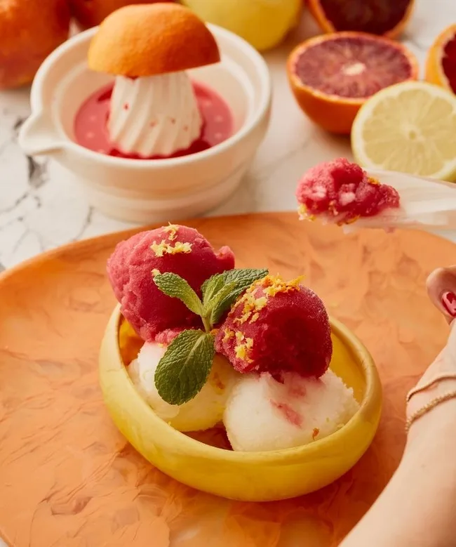 Colorful sorbet scoops in a yellow bowl with mint garnish, surrounded by citrus fruits and juice squeezer.