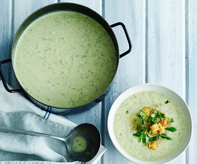 Creamy soup with herbs in a pot and a bowl, garnished with croutons and herbs on a light wooden surface.