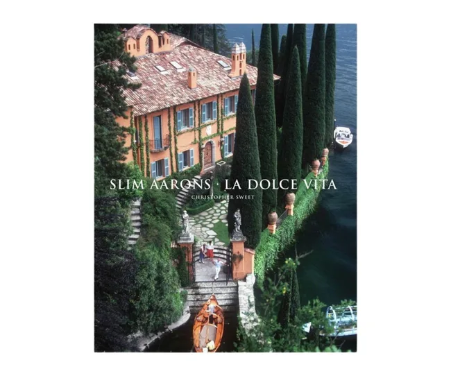 Cover of "La Dolce Vita" by Slim Aarons, showing a villa by a lake with lush greenery and a wooden boat.