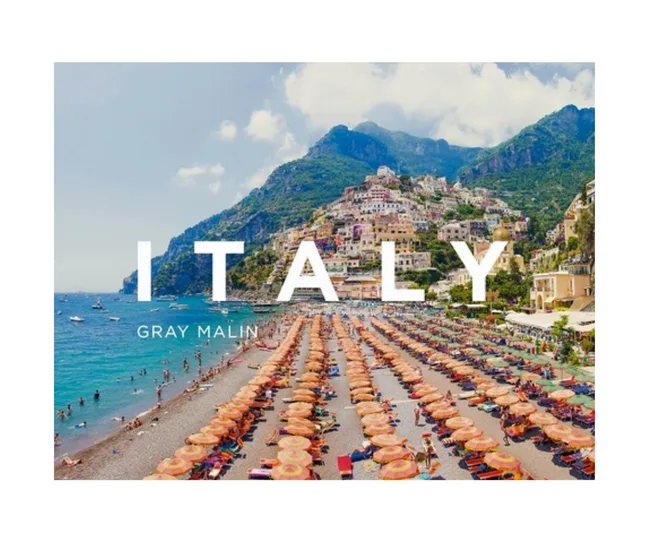 Beach umbrellas line a sunny shore in Positano, Italy, with colorful hillside buildings. Text: "ITALY" and "GRAY MALIN."