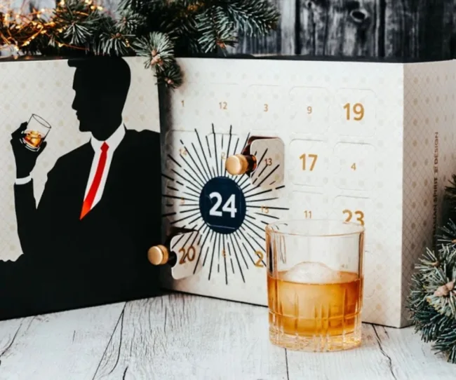 Advent calendar with numbers, glass of whiskey, and silhouette holding a drink, surrounded by pine branches.