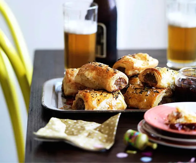 Sausage rolls on a tray with herbs, served with beer and condiments on a table.