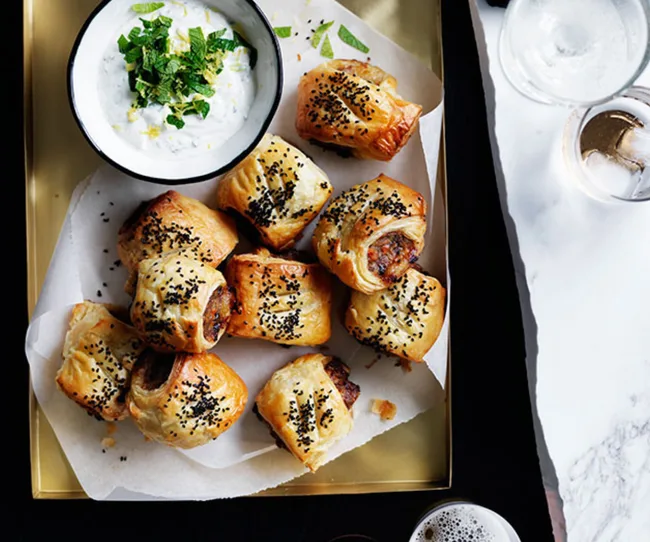 Mini sausage rolls topped with black sesame seeds, served with a mint yogurt dip on a golden tray.