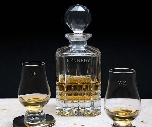 Cut glass decanter labeled "Kennedy" with two whiskey glasses marked "CK" and "WK," filled with amber liquid.