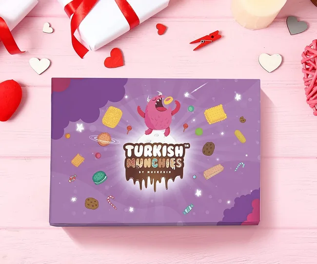 Gift box with "Turkish Munchies by Muezoom" on a pink table surrounded by red and white heart decorations.
