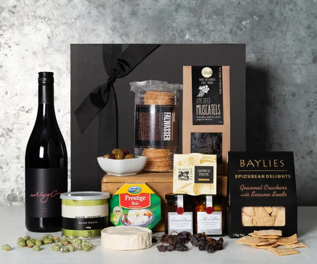 Gift box with red wine, cheese, crackers, quince paste, muscatels, olives, and wasabi peanuts on a marble surface.