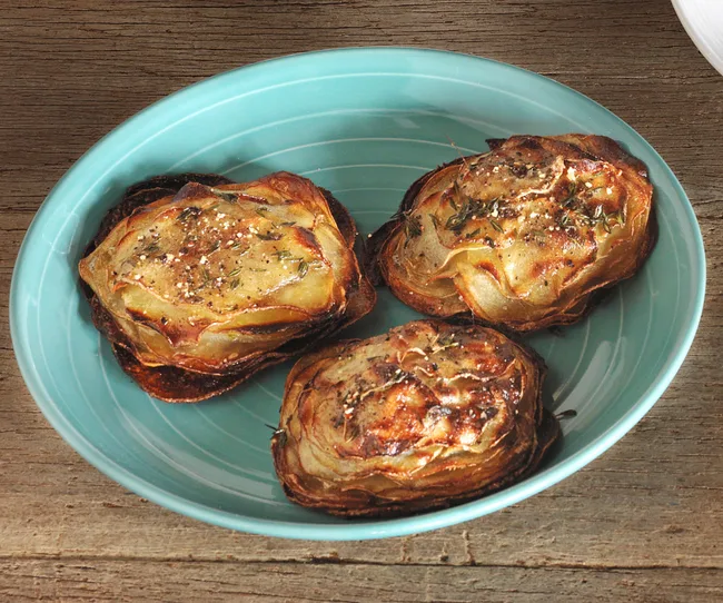 Three roasted potato stacks seasoned with herbs and spices in a light blue dish on a wooden surface.