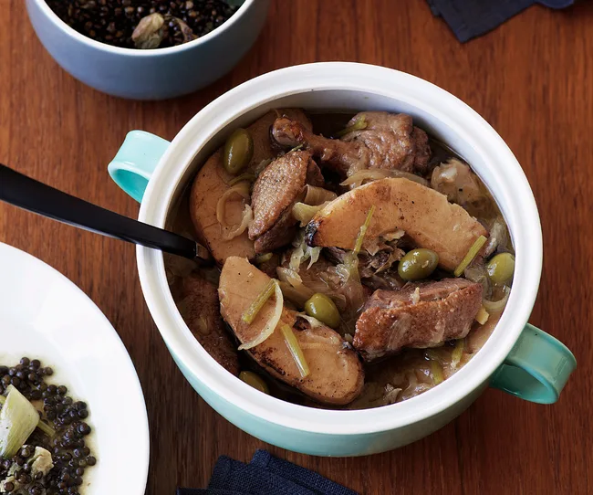 Duck tagine with apples, olives, and onions in a blue pot on a wooden table.