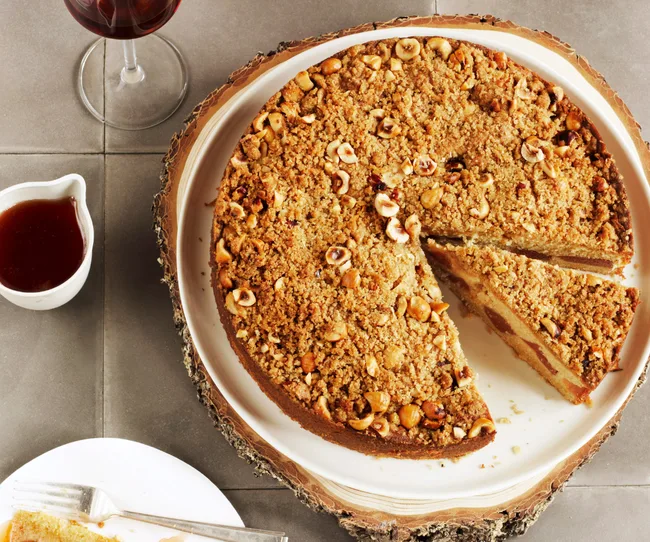 Apple hazelnut cake on a wooden platter, with a slice removed. A glass of red wine and sauce on the side.