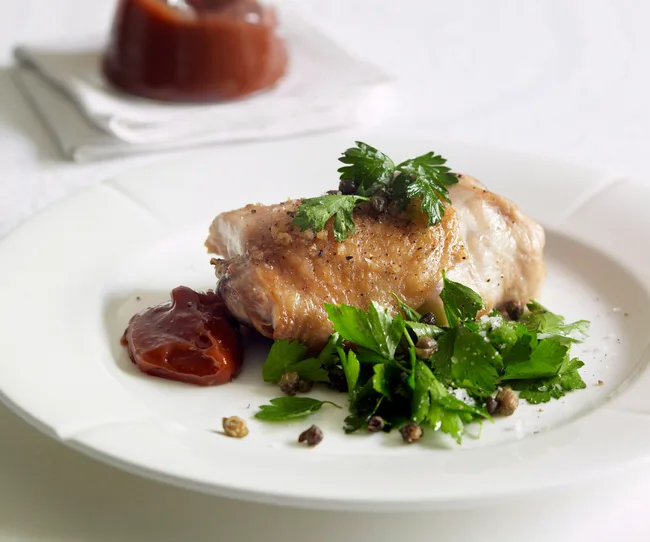 Roasted chicken with herbs and capers on a white plate, served with a side of sauce.