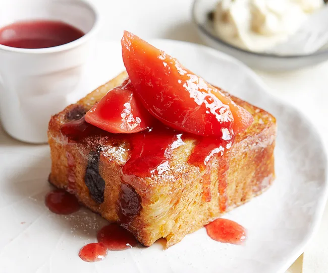 French toast topped with poached quince slices and drizzled with syrup on a white plate, with sauce and cream in the background.