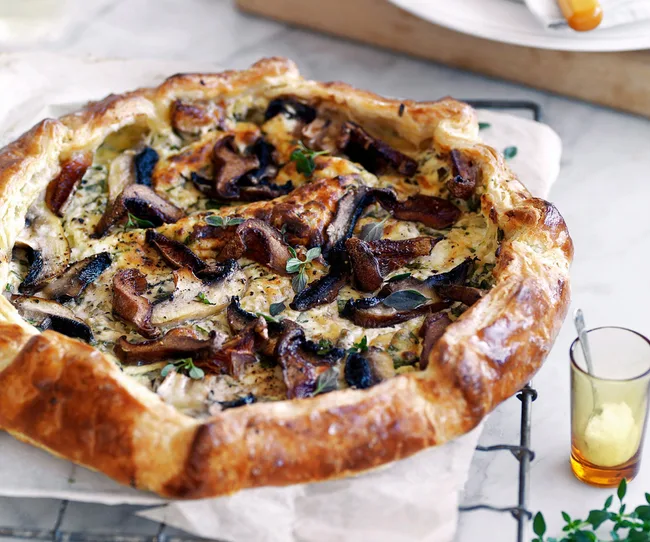 Rustic mushroom and cheese tart on a wire rack, with herbs and a small glass of seasoning beside it.