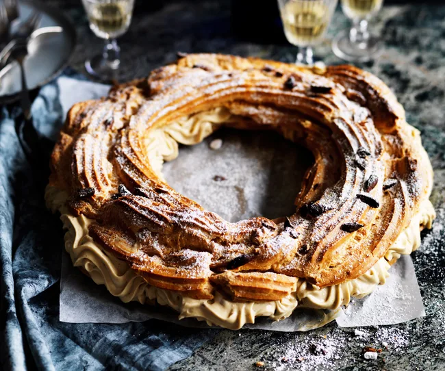 Golden Paris-Brest pastry with cream filling, topped with powdered sugar and nuts, on a rustic table setting.