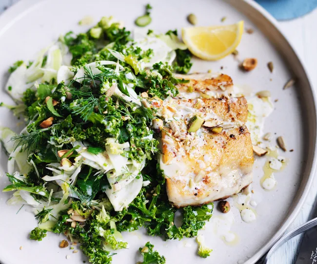 Grilled fish fillet with kale salad, seeds, and a lemon wedge on a white plate.