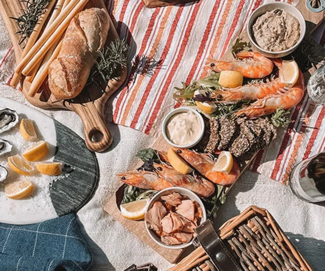 Seafood picnic spread with prawns, oysters, bread, pate, and dips on a striped cloth, with a wicker basket and drink.