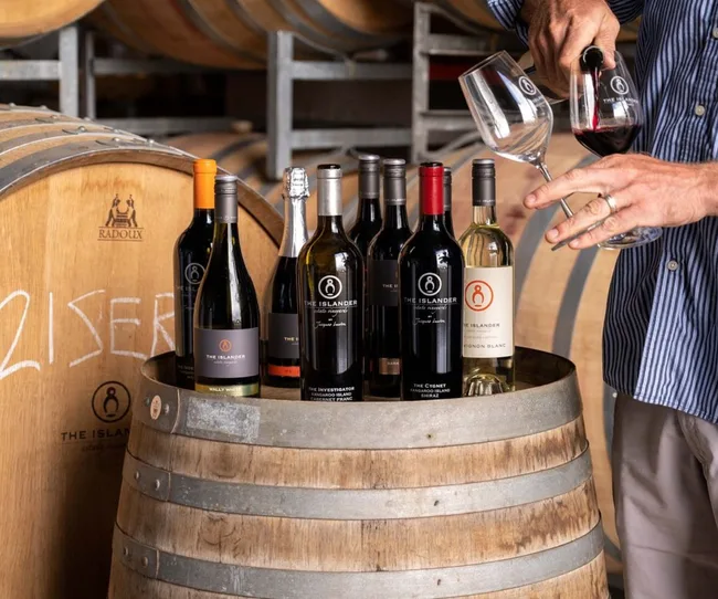 Man pouring red wine from a bottle labeled "The Islander" into a glass on a wine barrel, surrounded by various bottles.