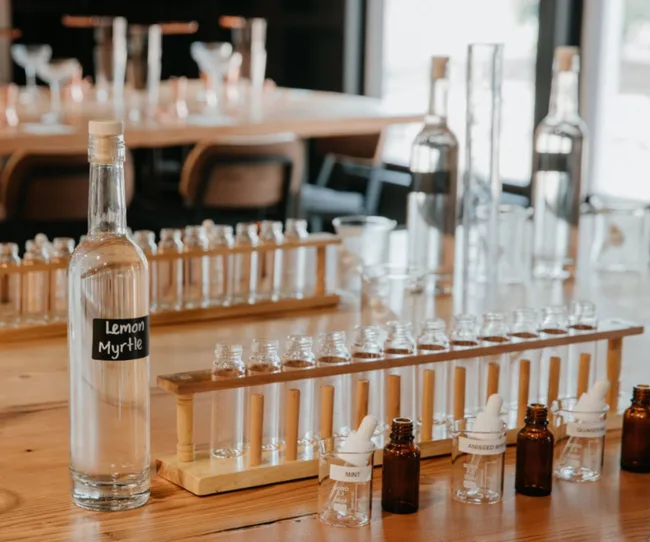 Bottles and test tubes on a wooden table, with a "Lemon Myrtle" labeled bottle centered among glassware.