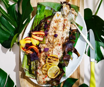 Aerial view of a barbecued coral trout wrapped partially in a banana leaf with grilled lemons and carrot escovitch.