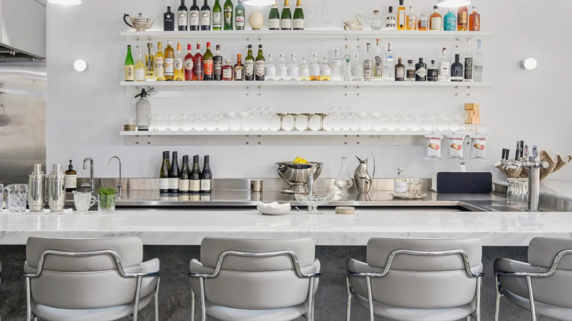 A photograph of the ice-toned marble bar and stools at Institut Polaire. Behind it, shelves stacked with alcohol and coupes. 