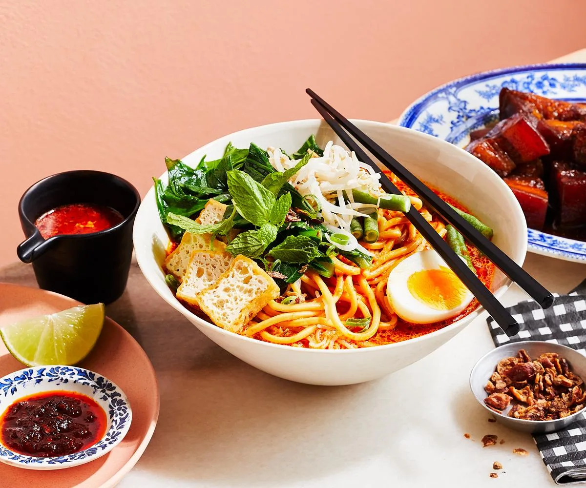 A bowl of curry laksa, with noodles, sliced fried tofu, a halved hard-boiled egg and a sprig of mint, with a pair of black chopsticks laid on top of the bowl.