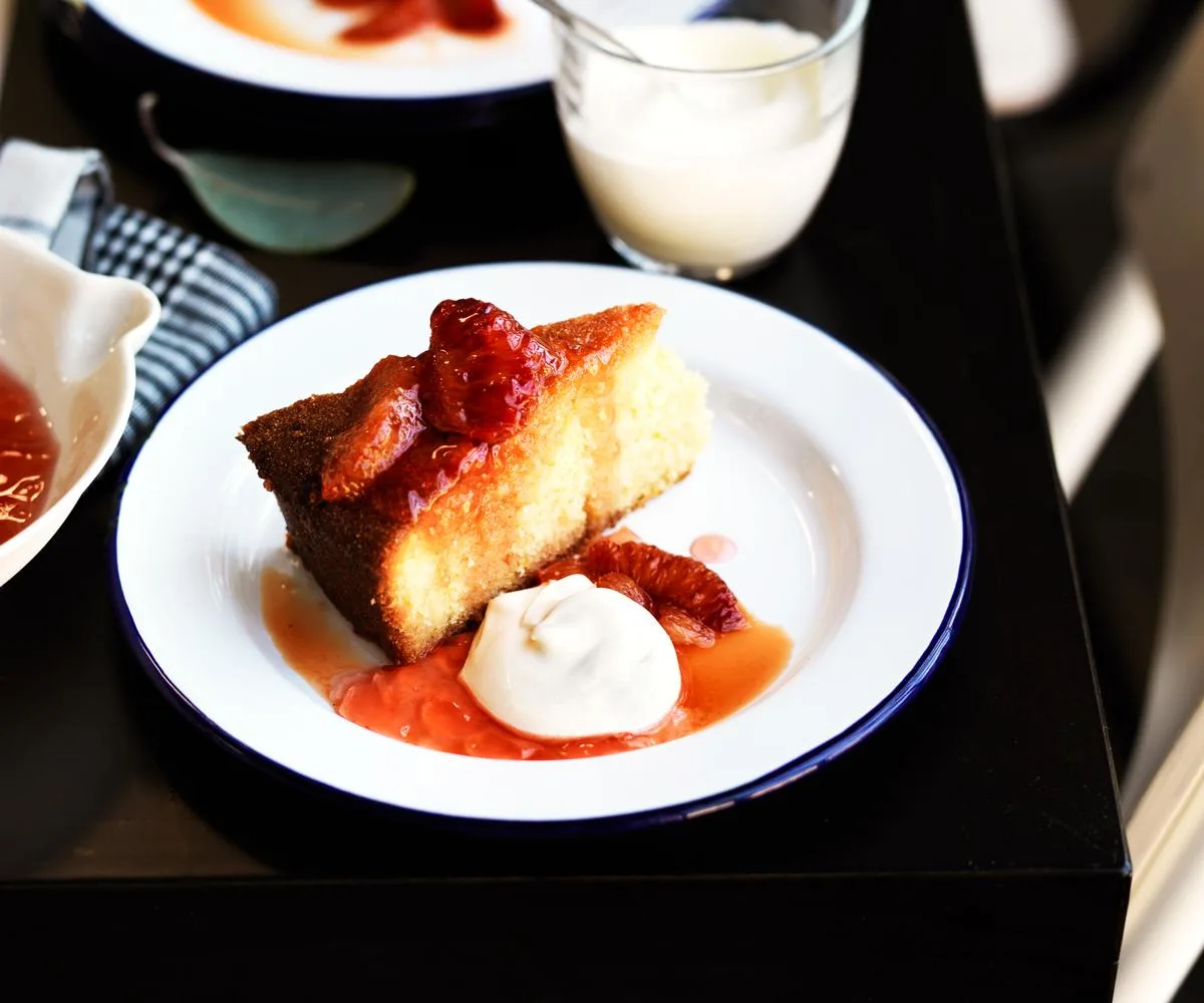 A round cake with a slice cut out of it, topped with segments of blood orange on a white round plate