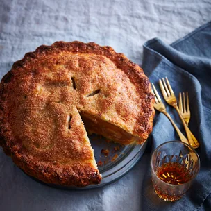 A round, crusty apple pie with a slice taken out of it, sitting on a light-blue linen tablecloth, with golden forks and a tumbler of amber-hued liquid to the side.