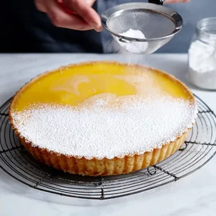 A whole lemon tart on a black wire rack being dusted with icing sugar.